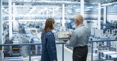 İki çok ırklı mühendis bir platformda duruyor, bir elektronik fabrikasında dizüstü bilgisayar kullanıyorlar. Makineler bakım ve uzmanlardan geçiyor.
