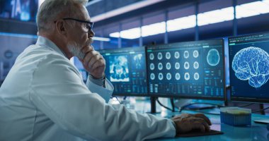 Kıdemli Erkek Nörobilimci, Tıbbi Araştırma Merkezi 'ndeki MRI taramalarını analiz etmek için Masaüstü Bilgisayarını kullanıyor. Beyaz Doktor CT 'yi inceliyor.