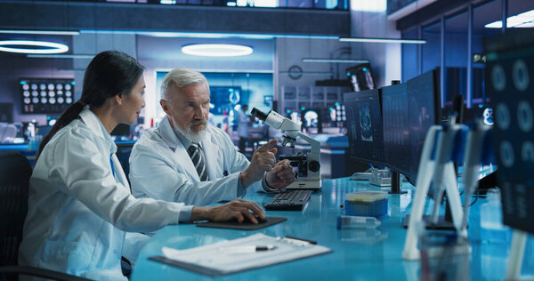 Центр медицинских исследований в области биотехнологий: женщина-азиатский нейробиолог, использующая настольный компьютер и разговаривающая с мужчиной-хирургом с помощью микроскопа