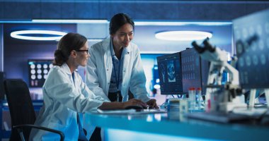 Tıbbi Araştırma Merkezi 'nde konuşan Asyalı ve Kafkas kadın doktorlar beyin tomografisini incelemek için masaüstü bilgisayarını kullanıyorlar. Cerrah ve Nörobilimci