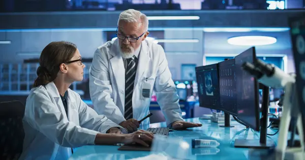 stock image Biotechnology Research Laboratory: Two Caucasian Doctors Discussing CT Brain Scan On Computer. Focus Shifting to Female Asian Scientist Working With