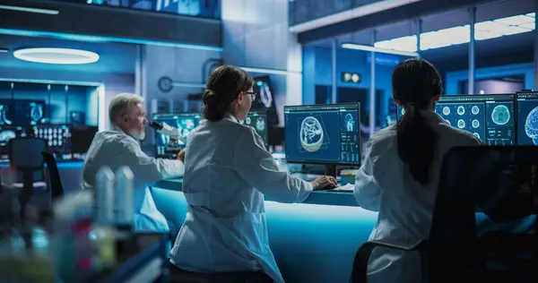 Stock image Back View Of Diverse Scientists Working In Medical Research Center. Male Doctor Using Microscope And His Female Colleagues Looking At CT Brain Scans