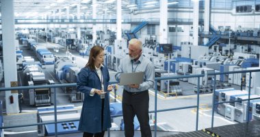 İki çok ırklı mühendis bir platformda duruyor, bir elektronik fabrikasında dizüstü bilgisayar kullanıyorlar. Makineler bakım ve uzmanlardan geçiyor.