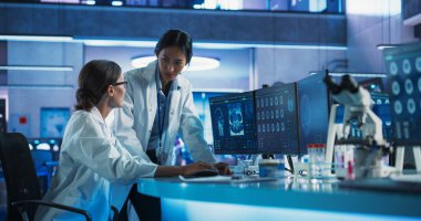 Tıbbi Araştırma Merkezi 'nde konuşan Asyalı ve Kafkas kadın doktorlar ve MRI beyin taramalarını analiz etmek için masaüstü bilgisayarı kullanıyorlar. Cerrah ve