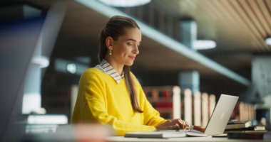 Sarı Jumper 'lı güzel beyaz kadın öğrenci dizüstü bilgisayarında okul tezi üzerinde çalışıyor. Çevrimiçi Çalışan Genç Zeki Kadın