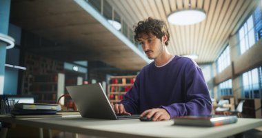 Modern Kütüphanede Öğrenci Çalışması. Genç Düşünen ve Sorunları Çözen Okul Egzersizleri. Üniversitede Çalışmak için Erkek Dizüstü Bilgisayarı Kullanıyor