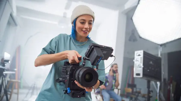 stock image Young Female Cinematographer With Blue Hair Adjusts Professional Camera In Studio, Showcasing Teamwork In Creative Film Production Environment, With