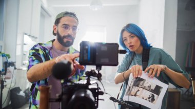 Parlak bir stüdyoda, genç beyaz bir erkek ve mavi saçlı bir kadın dijital kamerada gözden geçiriliyor, etrafı film yapımı ekipmanlarla çevrili, somutlaştırılmış