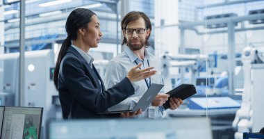 Teknoloji fabrikasında çalışan Endüstriyel Mühendisler. Kafkasyalı Adam ve Asyalı Kadın Laptop ve Tablet Bilgisayar Kullanıyor ve Konuşuyorlar