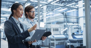 İki Çoklu Etnik Mühendis Bir Elektronik Fabrikasında Dizüstü bilgisayar ve Tablet Bilgisayar Kullanarak Ofiste Duruyor. Makineler bakım aşamasında ve...