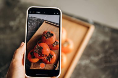 Kadın, gri arka planda akıllı telefonlu hurma meyvesinin fotoğrafını çekiyor.