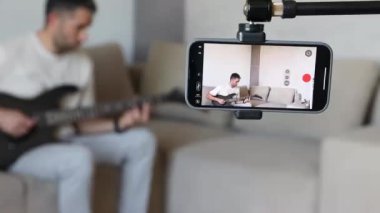 Beyaz bir adam elektro gitarla çalar ve çaldıklarını akıllı bir telefona kaydeder. Video dersleri, eğitim, blog yazma kavramı