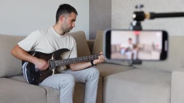 Beyaz bir adam elektro gitarla çalar ve çaldıklarını akıllı bir telefona kaydeder. Video dersleri, eğitim, blog yazma kavramı