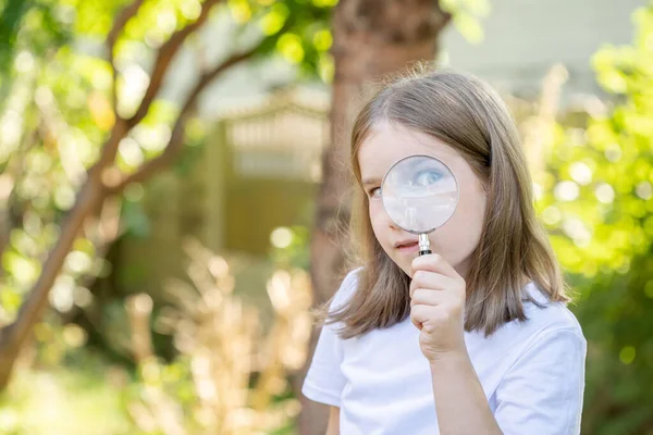一个好奇聪明的小学生 一个女孩用放大镜看着摄像机 手里拿着一个百叶窗 一只眼睛闭上 一只眼睛看得见 一个复制的空间 模糊的背景 年轻探险家 — 图库照片