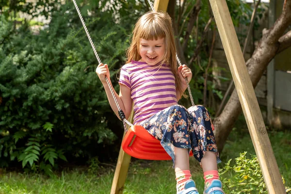 Happy girl, elementary school age child sitting swinging on an simple outdoor swing in the backyard, copy space, one person. Leisure activities, recreation concept, having fun outdoors, playground
