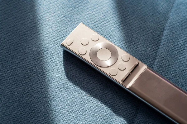 One single simple silver premium smart TV remote control laying on a sofa, couch, object in the sun, detail, closeup, nobody Watching TV symbol, digital recreation and leisure at home abstract concept