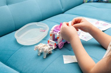 Genç kız, yumuşak mavi kanepe, odaklanmış renkli lego tuğlaları. El kitabından montaj kılavuzunu takip eden parçaları birleştiren eller, yakın plan.