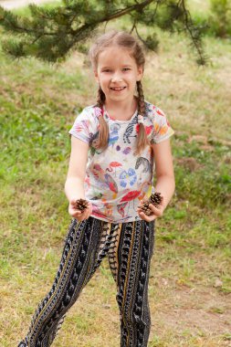 Ellerinde kozalak tutan genç bir kız, yaz ormanlarında gülümseyen çam kozalaklı bir çocuk, doğayı keşfeden çocuklarla bağ kuran doğa keşfi, açık hava eğlencesi.