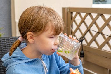 Girl drinking fresh water from a glass outdoors, healthy hydration, child enjoying beverage, casual dining, patio setting, young girl in blue hoodie, focus on healthy lifestyle and relaxation clipart