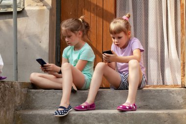 Akıllı telefonlar kullanarak beton basamaklarda oturan iki genç kız, açık havada cep telefonu ekranları, modern çocukluk, sosyal medya veya oyun bağımlılığı konsepti, çevrimiçi çocuk güvenliği