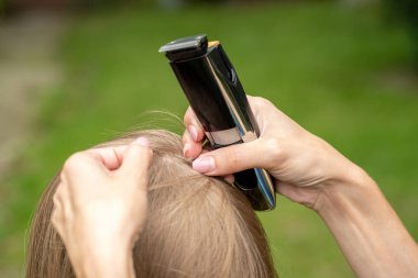 Çocuğun kafa derisini inceleyen, açık havada elektrikli makasla kızın saçını kesen, annesinin saç biti kontrolü yapan ve saç kesme makinesini yakın mesafeden tutan bir anne.