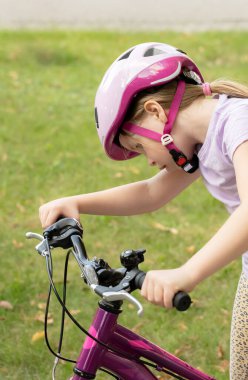 Pembe bisiklet kasklı okul kızı parkta sürmeden önce mor bisiklet gidonları ve frenleri kontrol ediyor, çocuk güvenlik denetimi sorumlu ekipman ayarlaması, yaz günü, dikey kapanış.