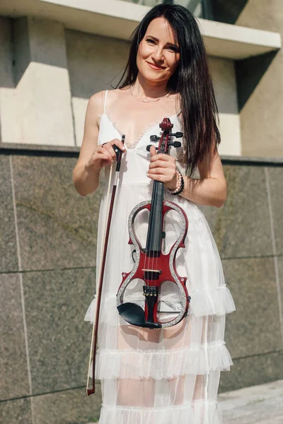 ドレスに暗い髪の女性アーティストが彼女の手に木製のコンサート電気バイオリンを保持しています — ストック写真