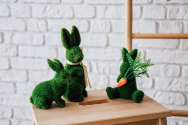 Üç yeşil yosun tavşanı ahşap bir masanın etrafında toplanıyor ve havuçla neşeli bir bahar teması sergiliyorlar..