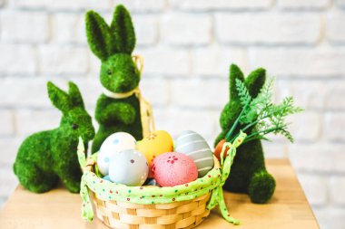 Pastel renkli Paskalya yumurtalarıyla dolu bir sepeti çevreleyen yeşil malzemeden yapılmış dekoratif tavşanlar. Ayar aydınlık ve neşeli..