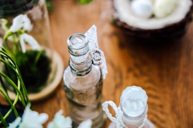 Dantelle süslenmiş iki cam şişe, sıcak bir iç mekanda bitkilerle ve dekoratif malzemelerle çevrili ahşap bir masaya oturur..