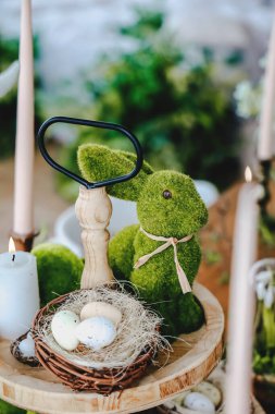 Yosun kaplı bir tavşan, yeşilliklerle çevrili ahşap bir masada Paskalya yumurtası sepetinin yanında duruyor..