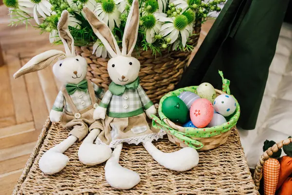 Taze çiçeklerle çevrili renkli yumurtalarla dolu bir sepetin yanında oturan iki sevimli tavşan baharın gelişini kutluyorlar..