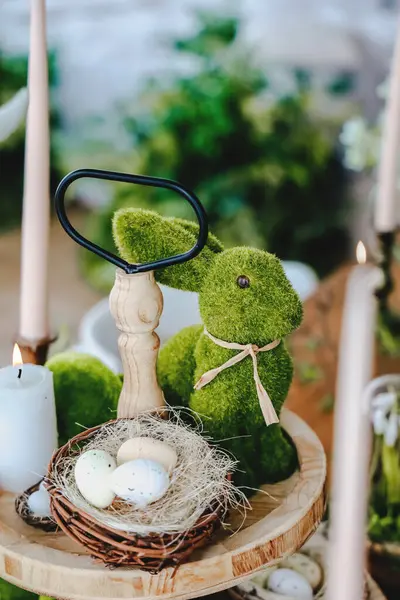 Yosun kaplı bir tavşan, yeşilliklerle çevrili ahşap bir masada Paskalya yumurtası sepetinin yanında duruyor..