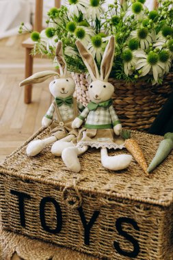 İki yumuşak pelüş tavşan, dekoratif havuçlar ve canlı çiçeklerle çevrili oyuncak bir sepetin üzerinde oturup büyüleyici bir atmosfer yaratırlar..