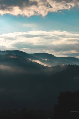 Yumuşak sabah ışığı bulutlu bir gökyüzünün altında yuvarlanan dağları aydınlatır. Huzurlu ve sakin bir atmosfer yaratır..
