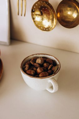 Kavrulmuş kahve çekirdekleri ile dolu seramik bir fincan, metalik çatal bıçakların yanında beyaz bir yüzeye oturmuş, rahat bir kafe atmosferi yaratıyor..