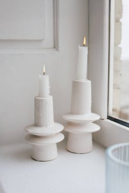 Minimalist tutucularda iki beyaz mum, sakin bir anda, parlak bir pencerenin kenarında yavaşça sıcak bir parıltı saçar..