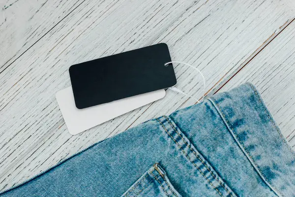 Bir kot pantolon, kırsal ahşap bir yüzeye dayanır. Beline iki fiyat etiketi iliştirilmiş, yeni bir alımı vurguluyor..