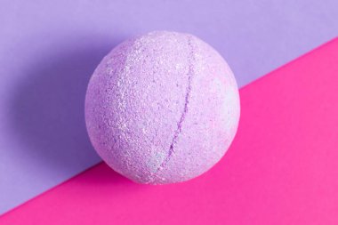 Yuvarlak mor bir banyo bombası, parlak pembe ve mor bir arka planda durur. Spa günleri ve kişisel bakım için mükemmeldir..