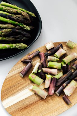 Taze doğranmış kuşkonmaz, ahşap bir kesim tahtasının üzerinde tüm kuşkonmaz saplarının yanında canlı yeşil ve mor renkler sergiliyor..