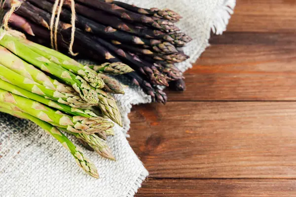 Yeşil ve mor kuşkonmaz demetleri ahşap bir masadaki desenli kumaşa dizilir ve canlı renkleri ve taze görünüşleri sergilenir..