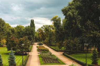 Bereketli ağaçların ve bulutlu bir gökyüzünün altında canlı çiçeklerle çevrili bir şehir parkından geçen huzurlu bir patika..