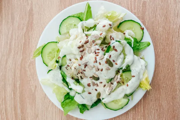 Kıtır kıtır salatalıklı canlı bir salata ve üzerine kremalı sos ve tohum serpiştirilmiş çıtır marul. Tahta bir masada sağlıklı bir yemeğin tadını çıkarıyorum..