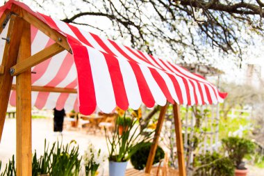 Canlı kırmızı ve beyaz çizgili tente, çiçek ve bitkilerle dolu verimli bir bahçe alanında ahşap masaların üzerine gölge sağlar..