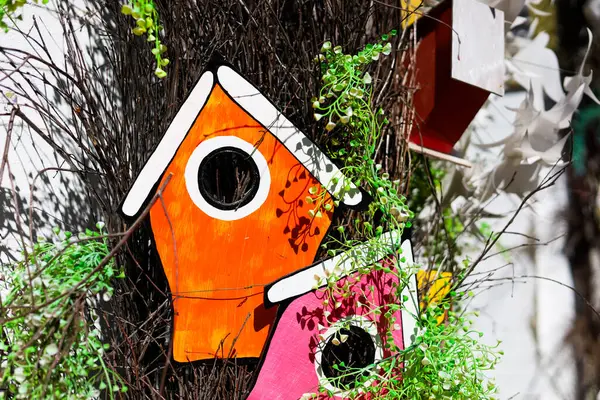 Turuncu ve pembe boyalı iki kuş evi yeşilliklerle çevrili bir ağaçta asılı, canlı bir bahçe ortamı yaratıyor..