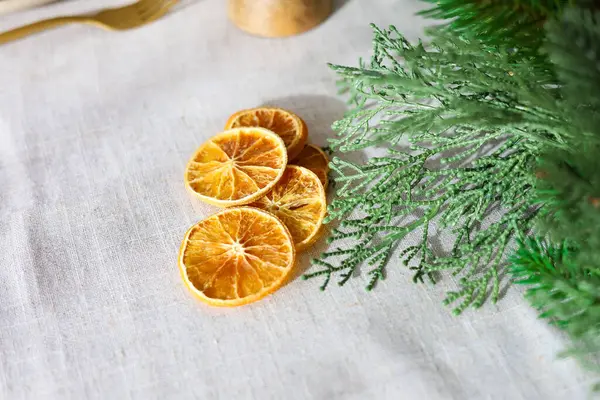 Kurutulmuş portakal dilimleri keten bir masa örtüsüne konur ve yanında yeşil yapraklar doğal bir dekor oluşturur..