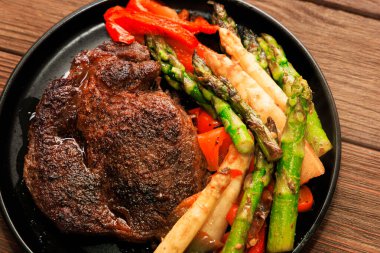 Sulu ızgara biftek, kuşkonmaz ve dolmalık biber de dahil olmak üzere rengarenk kavrulmuş sebzelerin yanında siyah bir tabağa konur..