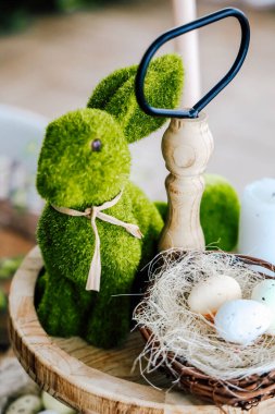 Yosun tutmuş canlı yeşil bir tavşan figürü pastel renkli yumurtalarla dolu bir yuvanın yanında duruyor ve doğal ahşap bir masa üzerinde ahşap bir alet..