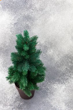 Küçük bir yapay yeşil ağaç gri bir yüzey üzerinde kahverengi bir tencerede duruyor. Tatil dekorasyonu için ideal yaprakları sergiliyor..