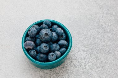 Taze toplanmış yaban mersinleriyle dolu turkuaz bir kase açık renkli bir yüzeye oturmuş canlı renklerini ve tazeliklerini sergiliyor..